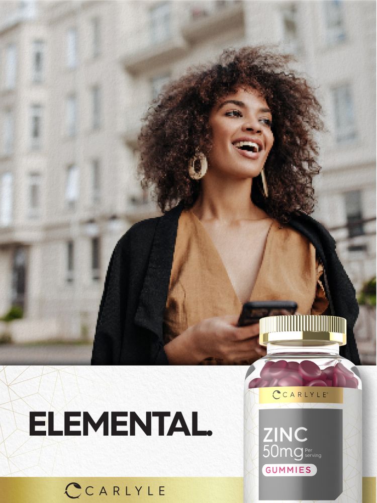 A young adult woman in the city smiles and holds a phone, along with a bottle of Zinc 50 mg Gummies.