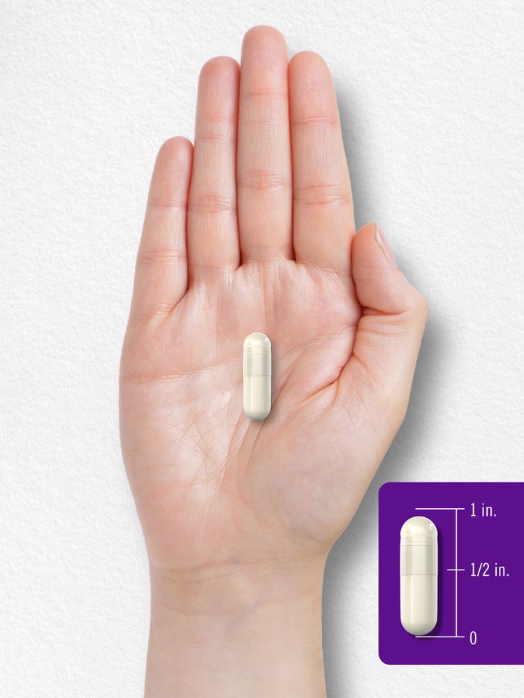 A light-colored capsule in an open hand, with a scale showing it measures 1 inch, marked at 1/2 inch and 0 inches.