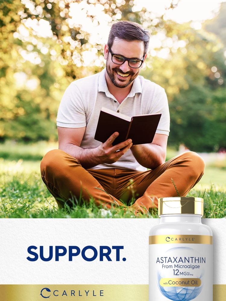 A man sits on grass reading a book with a Carlyle Astaxanthin bottle beside him.