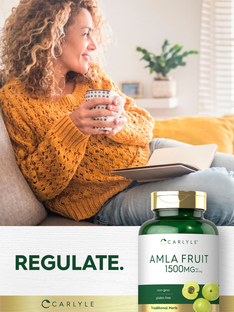 Adult woman sits on a sofa, holding a mug and book, smiling while looking away, next to a Carlyle Amla Fruit bottle.
